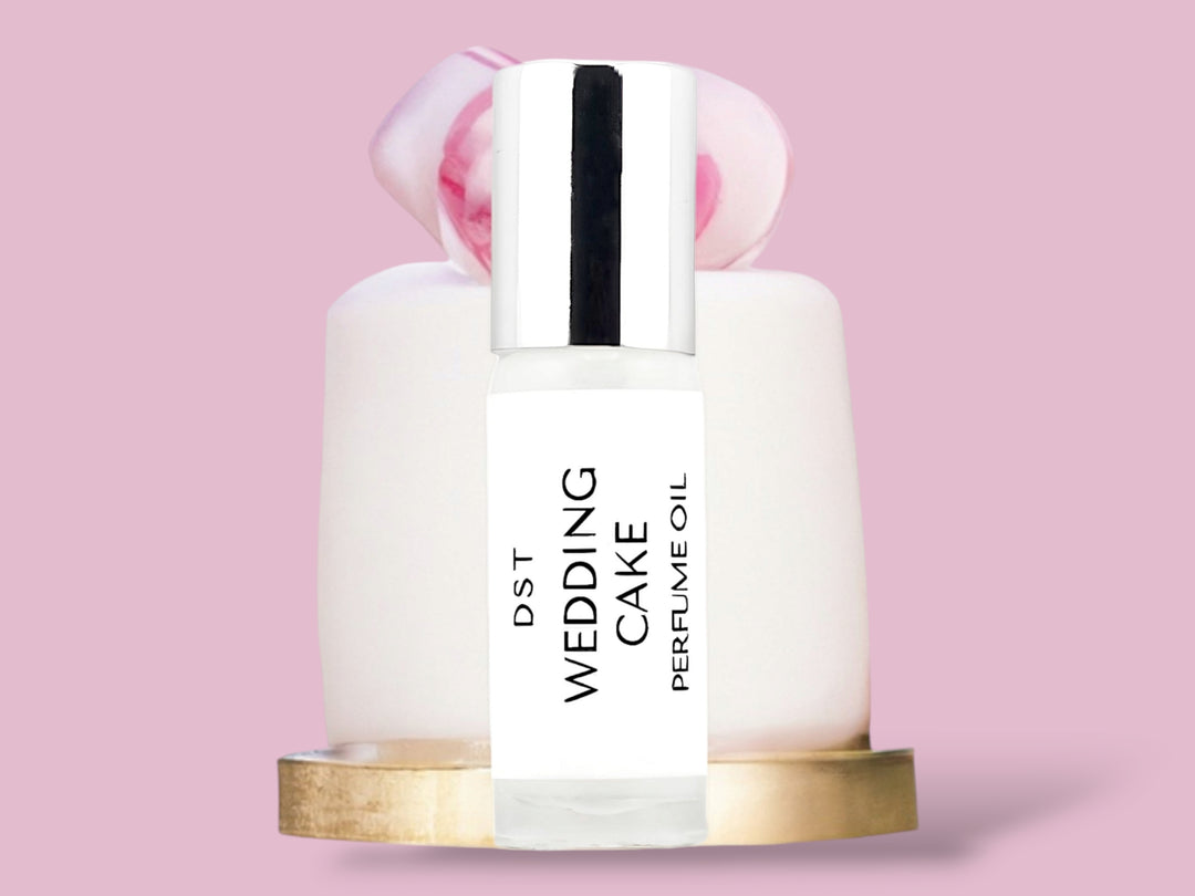 5ml bottle of Wedding Cake perfume oil sitting in front of a simple one tiered white Wedding Cake on a gold plate with pink flower on top, against a pink background.