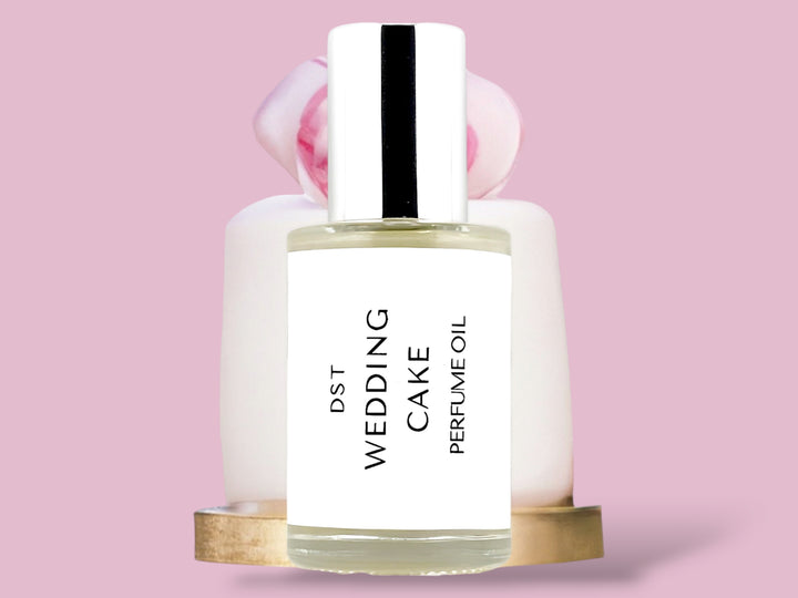 30ml bottle of Wedding Cake perfume oil sitting in front of a simple one tiered white Wedding Cake on a gold plate with pink flower on top, against a pink background.