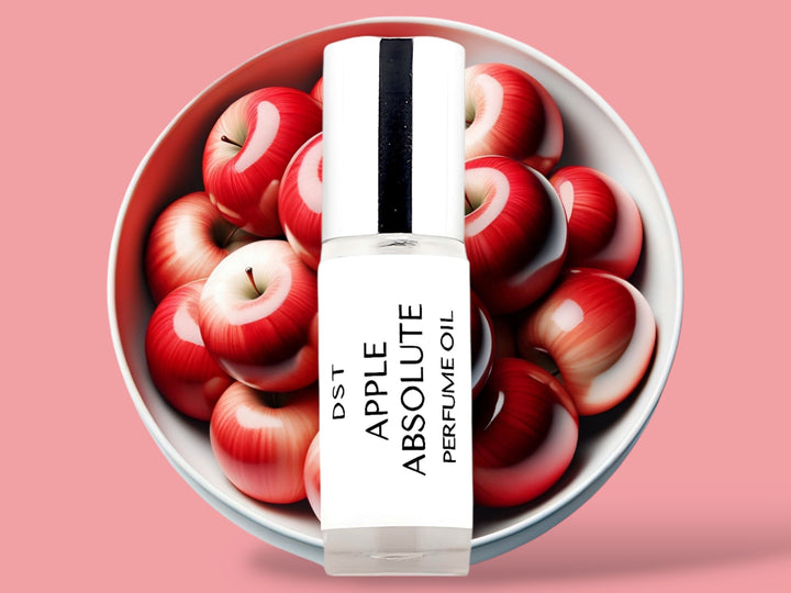 5ML bottle of Apple Absolute perfume oil sitting in front of a large white bowl filled with red apples against a light red background. 