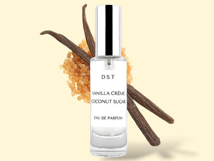 30ml bottle of Vanilla Creme and Coconut Sugar Parfum sitting in front of 2 large vanilla beans that are lying on top of a pile of brown sugar against a cream colored background. 