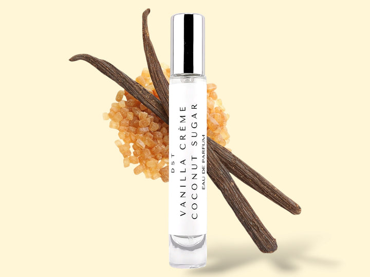 10ml bottle of Vanilla Creme and Coconut Sugar Parfum sitting in front of 2 large vanilla beans that are lying on top of a pile of brown sugar against a cream colored background. 