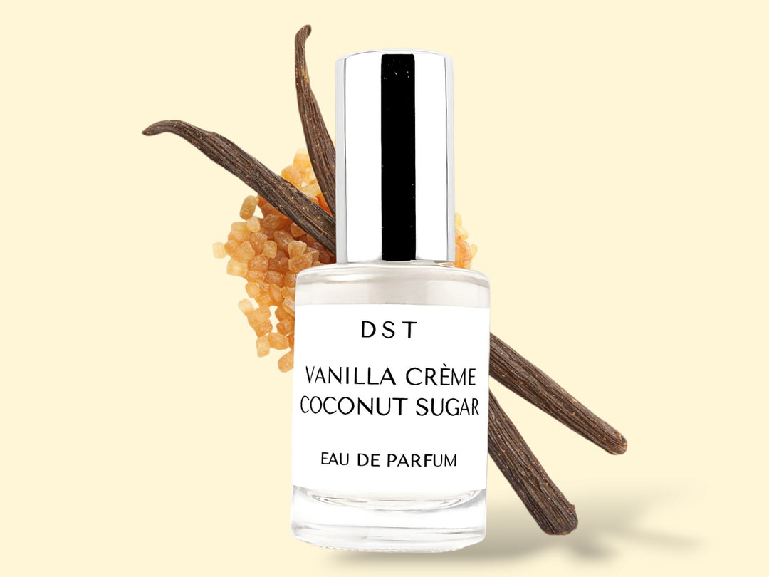 10ml bottle of Vanilla Creme and Coconut Sugar Parfum sitting in front of 2 large vanilla beans that are lying on top of a pile of brown sugar against a cream colored background. 