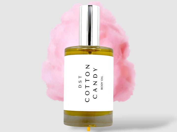 100ml bottle of Cotton Candy body oil sitting in front of a large Cotton Candy on a wooden stick against a grey background. 