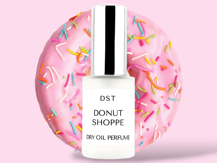 30ML bottle of Donut Shoppe Dry Oil Perfume spray sitting in front of a pink Frosted Donut with rainbow sprinkles against a pink background. 