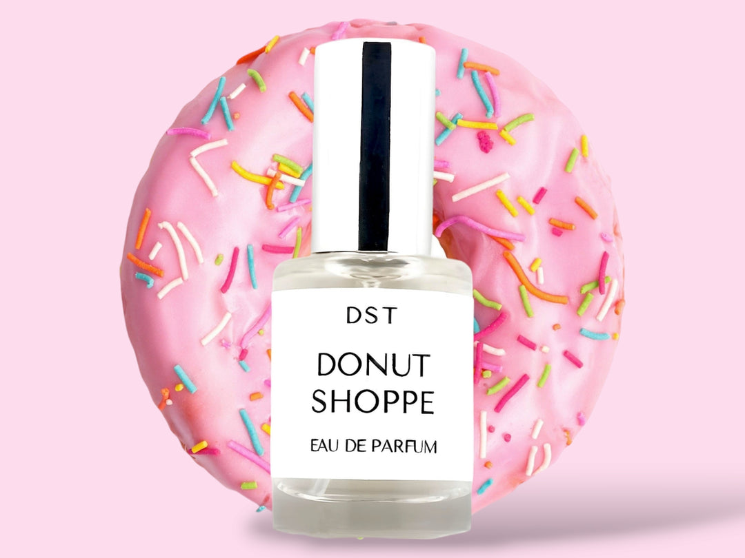 10ML bottle of Donut Shoppe Eau de Parfum sitting in front of a pink Frosted Donut with rainbow sprinkles against a pink background. 