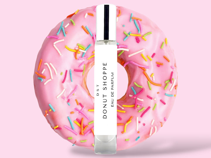 10ML bottle of Donut Shoppe Eau de Parfum sitting in front of a pink Frosted Donut with rainbow sprinkles against a pink background. 