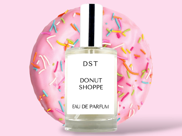 100ML bottle of Donut Shoppe Eau de Parfum sitting in front of a pink Frosted Donut with rainbow sprinkles against a pink background. 