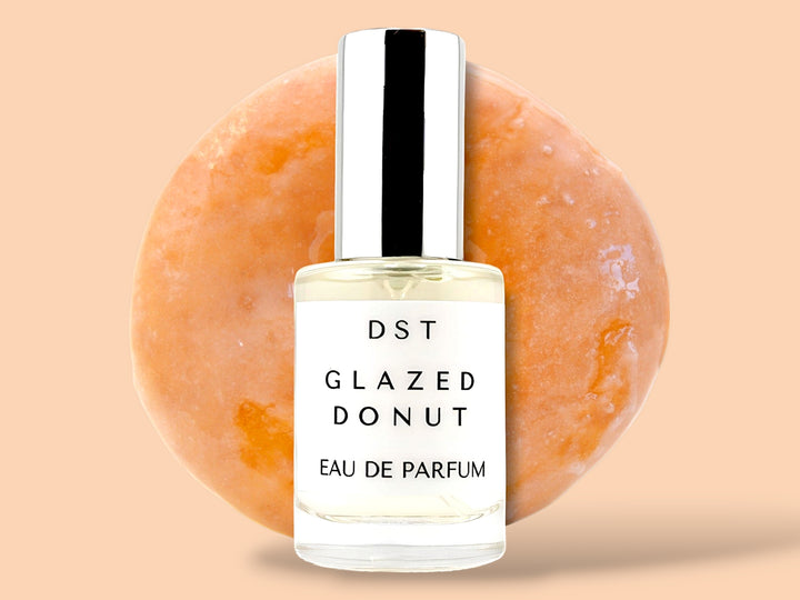 10ml bottle of glazed donut eau de parfum sitting in front of a large glazed donut on a cream colored background.