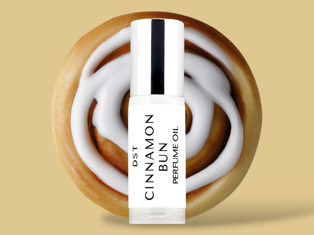 5ML bottle of Cinnamon Bun perfume oil sitting in front of a large Cinnamon Bun with white frosting against a tan background. 