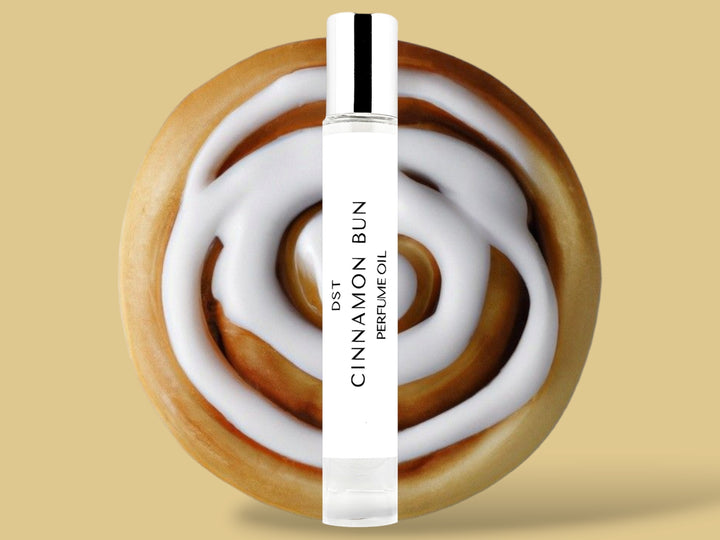 10ML bottle of Cinnamon Bun perfume oil sitting in front of a large Cinnamon Bun with white frosting against a tan background. 