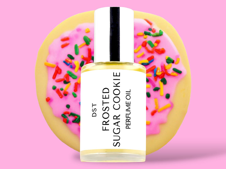 30ML bottle of Frosted Sugar Cookie perfume oil sitting in front of a large Sugar Cookie with pink frosting and rainbow sprinkles against a pink background. 