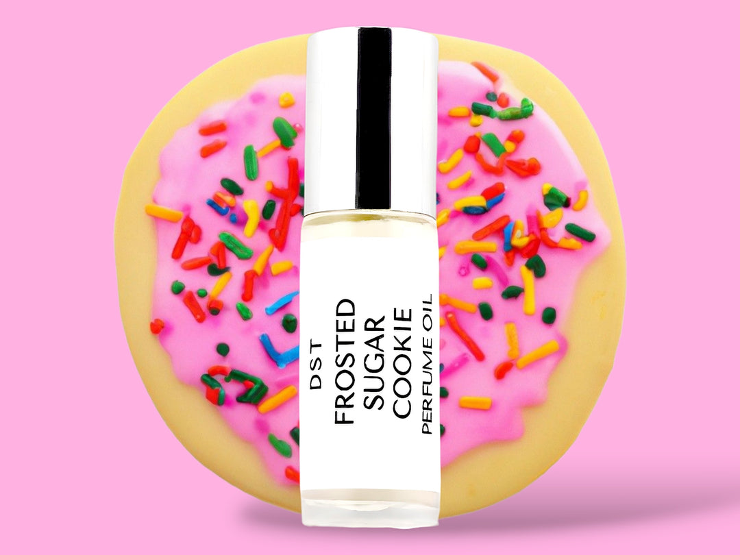 5ML bottle of Frosted Sugar Cookie perfume oil sitting in front of a large Sugar Cookie with pink frosting and rainbow sprinkles against a pink background. 