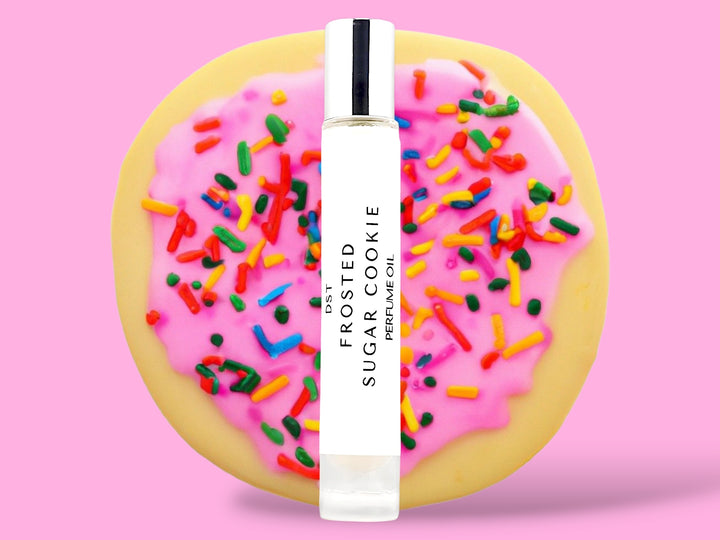 10ML bottle of Frosted Sugar Cookie perfume oil sitting in front of a large Sugar Cookie with pink frosting and rainbow sprinkles against a pink background. 