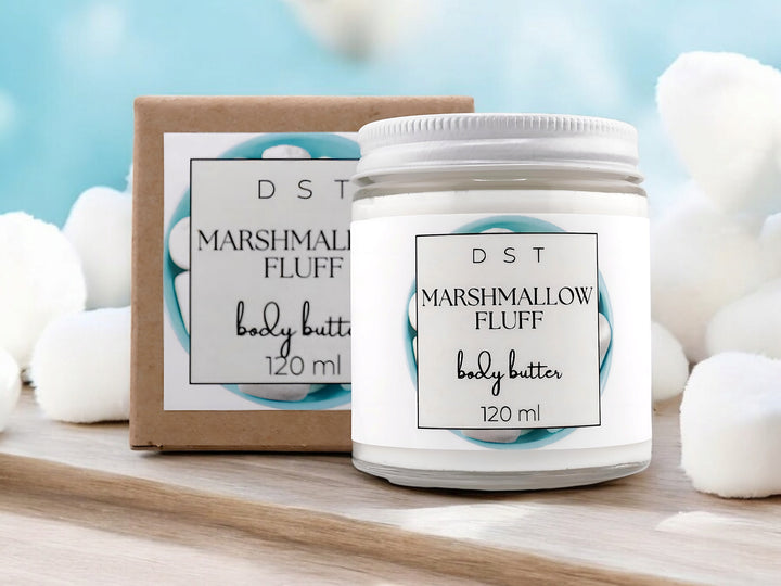 4oz jar of Marshmallow Fluff Body Butter and the box it is packaged in sitting on wood counter top with light blue background and white marshmallows.