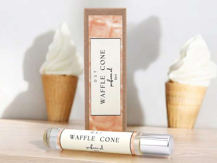 10ml bottle of waffle cone perfume oil and the box it is packaged in sitting on wood counter top with white background and 2 waffle cone filled with vanilla ice cream.