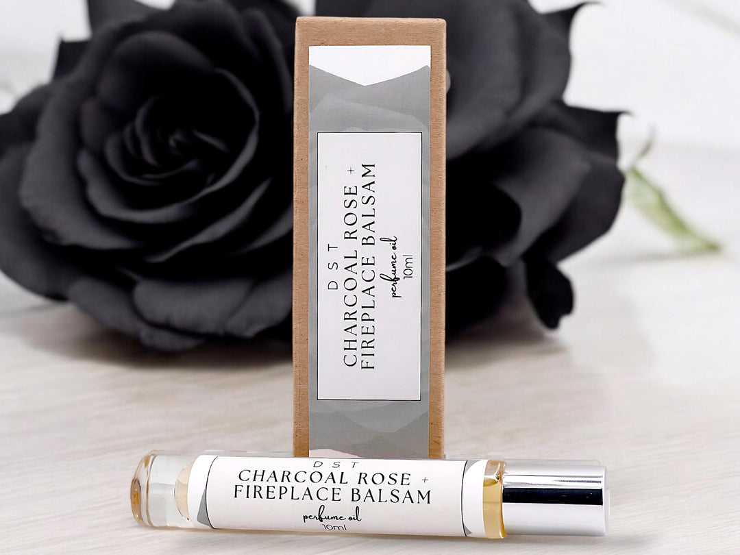 10ml bottle of Charcoal Rose and Fireplace Balsam Perfume oil and the box it is packaged in sitting in front of a large black rose on against a light grey background. 