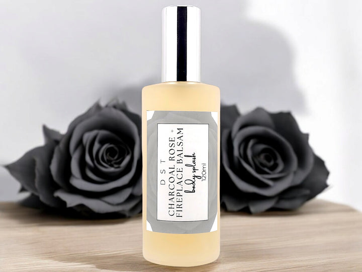 4oz bottle of Charcoal Rose and Fireplace Balsam body splash sitting on a wood counter top with black roses against a shadowy white background. 