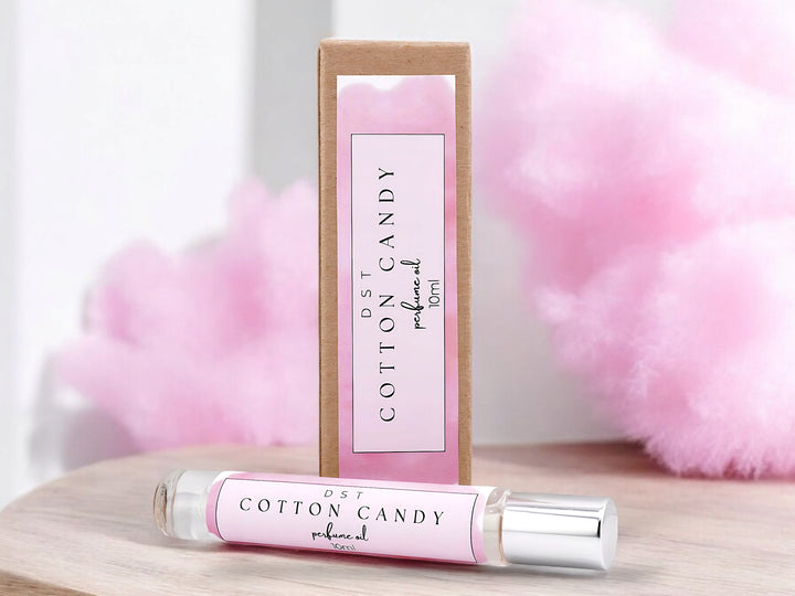 10ml bottle of Cotton Candy perfume oil and the box it is packaged in sitting  on wood counter top with fluffy pink cotton candy in the white shadowy background.