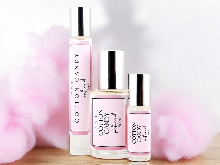 5, 10, and 30ml bottles of Cotton Candy perfume oil sitting wood counter top with fluffy pink cotton candy in the white shadowy background.