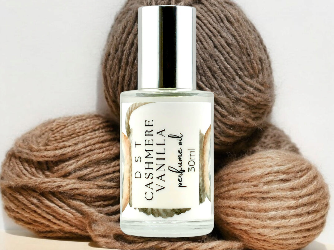 30ml bottle of Cashmere Vanilla perfume oil sitting in front of balls of tan colored yarn against a light gray background. 