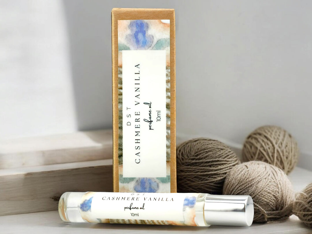 10ml bottle of Cashmere Vanilla perfume oil and the box it is packaged in sitting in front of balls of tan colored yarn against a light gray background.