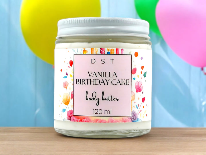 120ml jar of Vanilla Birthday Cake scented Body Butter sitting on wood counter top with yellow and pink balloons against a blue background.