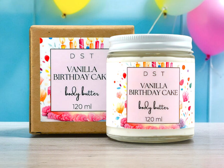 120ml jar of Vanilla Birthday Cake scented Body Butter and the box it is packaged in sitting on wood counter top with yellow and pink balloons against a blue background.