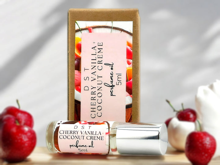 5ml bottle of Cherry Vanilla and Coconut Cream perfume oil and the box it is packaged in sitting on wood countertop with bright red cherries and whipped crem in the white background.