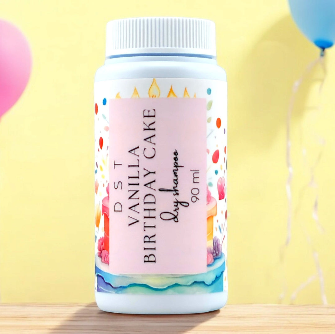 90ml Bottle of Vanilla Birthday Cake scented Dry Shampoo powder sitting on a wood counter top with pink and blue party balloons against a yellow background.