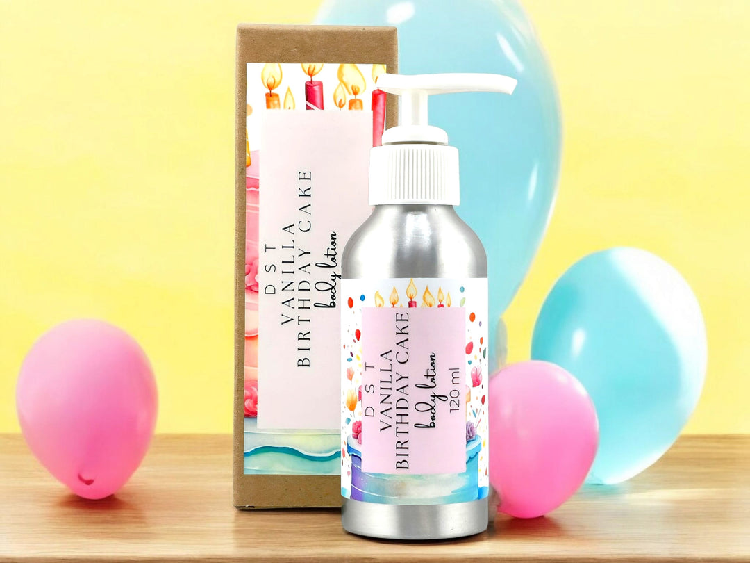 4oz Bottle of Vanilla Birthday Cake scented Body lotion and the box it is packaged in sitting on a wood counter top with pink, and blue party balloons against a yellow background.