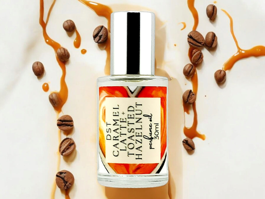 30ml bottle of Caramel Latte and Toasted Hazelnut Perfume Oil against Cream colored background surrounded by coffee beans and caramel drizzle. 