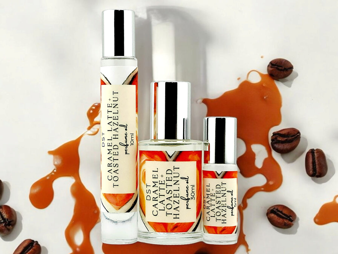 5, 10, and 30ml bottles of Caramel Latte and Toasted Hazelnut Perfume Oil against Cream colored background surrounded by coffee beans and caramel drizzle. 