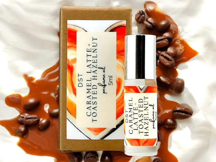 5ml bottle of Caramel Latte and Toasted Hazelnut Perfume Oil and the box it is packaged in against Cream colored background surrounded by coffee beans and caramel drizzle. 