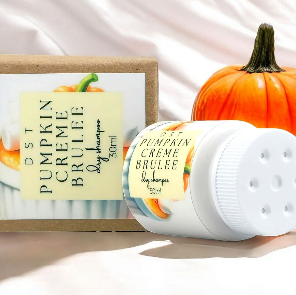 30ml bottle of Pumpkin Creme Brulée scented Dry Shampoo powder and the box it is packaged in sitting on a wood countertop with pumpkins in the ivory backdrop. 