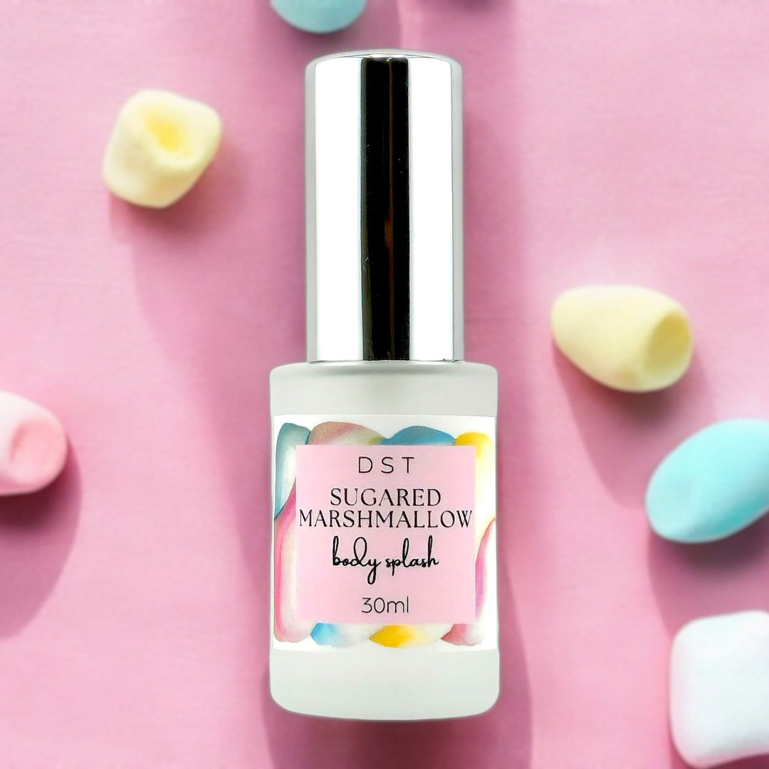 Thirty milliliter bottle of sugared marshmallow body splash sitting on a pink surface, surrounded by pastel blue , yellow and pink marshmallows.