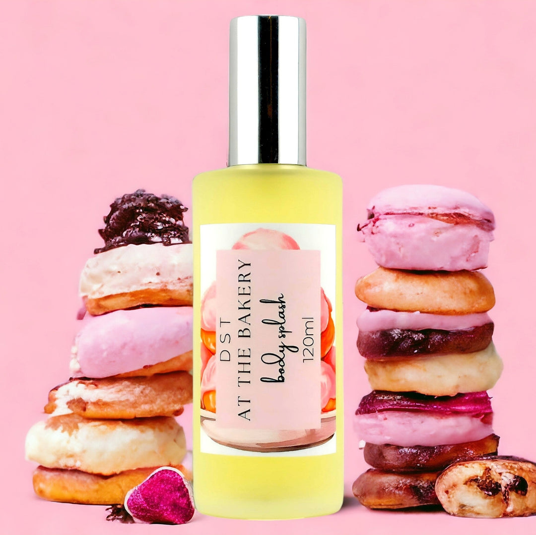 120ml bottle of AT the Bakery body splash against a pink shadowy background with color French pastries  stacked on each side of the bottle.