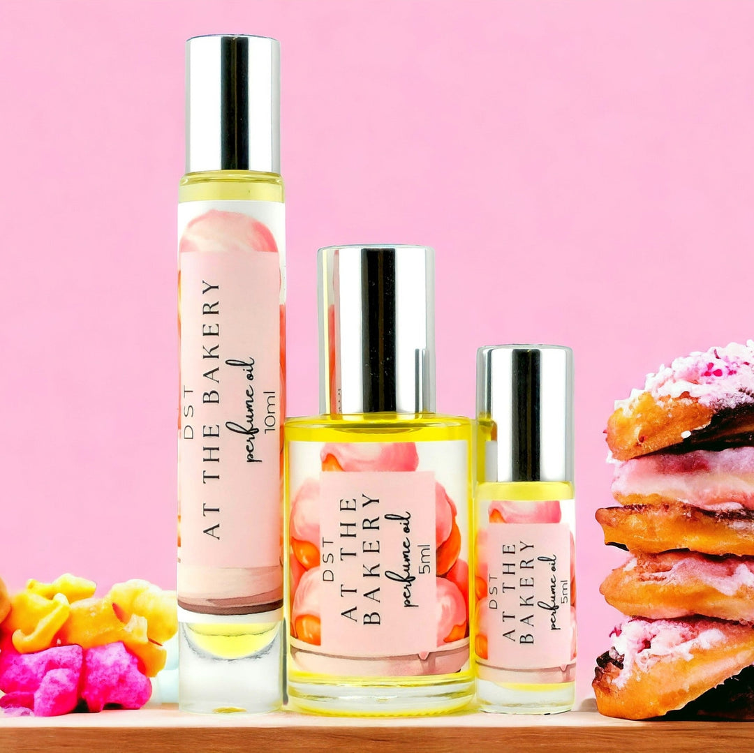 5, 10, and 30 ml bottles of AT the Bakery perfume oil against a pink shadowy background with color French pastries  stacked on each side of the bottle.