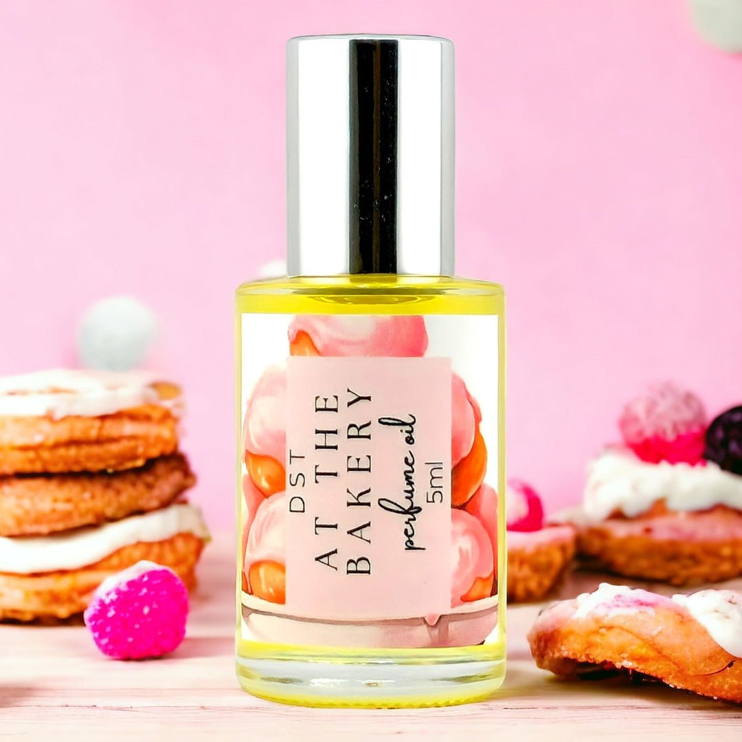 30ml bottle of AT the Bakery perfume oil against a pink shadowy background with color French pastries  stacked on each side of the bottle.