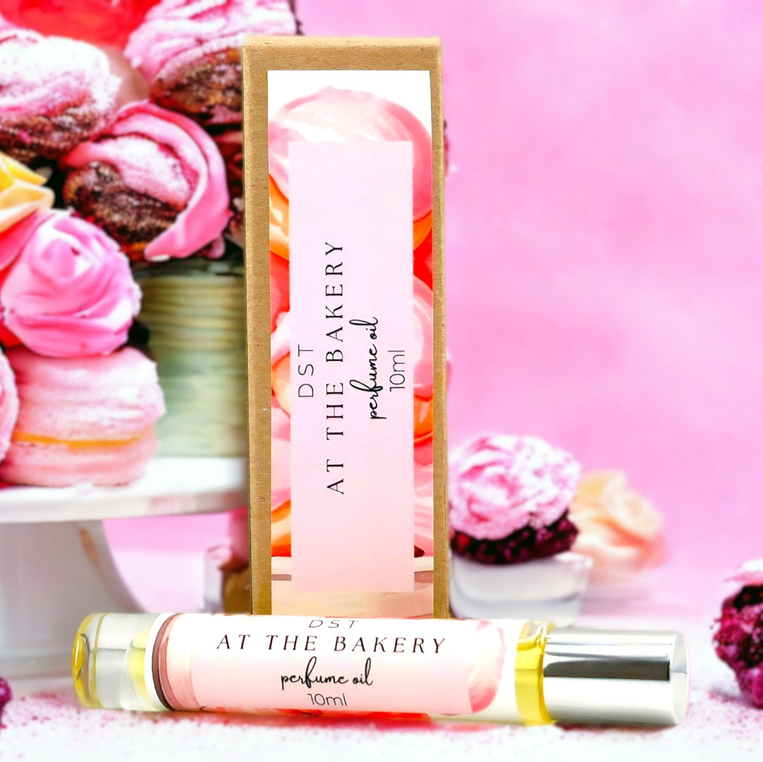 10ml bottle of AT the Bakery perfume oil and the box it is packaged in against a pink shadowy background with color French pastries  stacked on each side of the bottle.