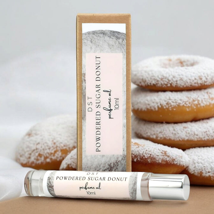 10ml bottle of Powdered sugar donut perfume oil and the box it is packaged in against a grey background with stacked powderedsugar donuts. 