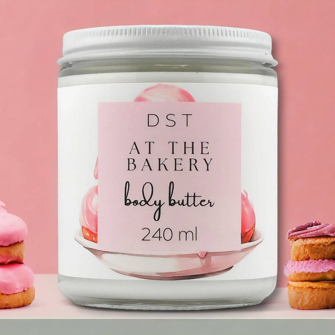 8oz JAR of AT the Bakery scented body butter against a pink background with colorful pastries stacked next to jar. 