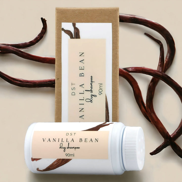 90ml bottle of Vanilla Bean scented Dry Shampoo powder and the box it is packaged in against ivory background with long vanilla beans. 