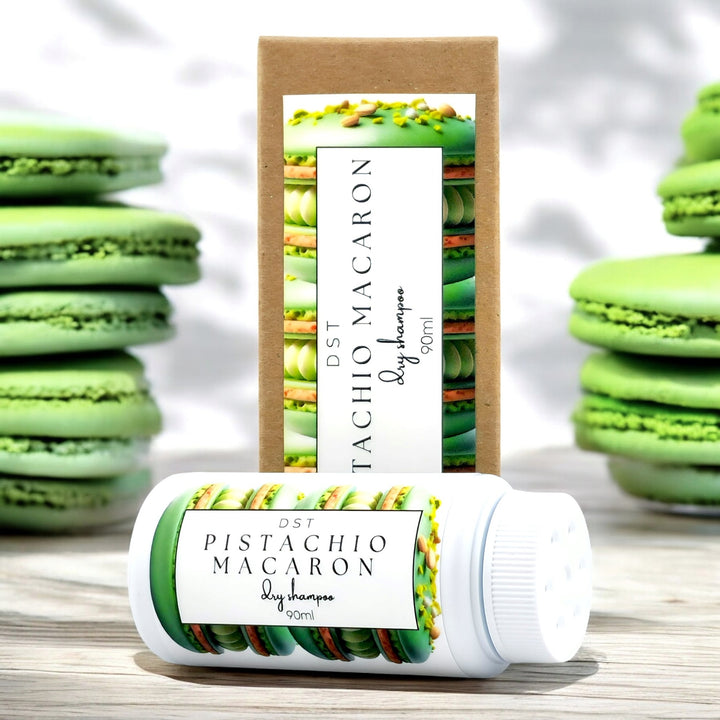 90ml bottle of Pistachio Macaron scented Dry Shampoo powder and the box it is packaged in sitting on a wood countertop with green French Macarons in the background.