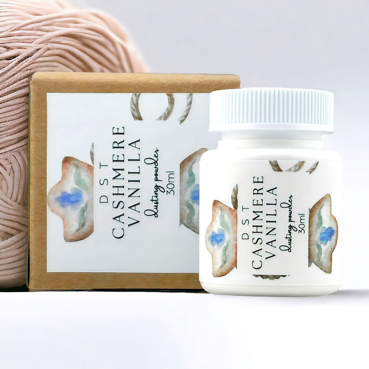 30ml bottle of Cashmere Vanilla scent body powder and the box it is packaged in against a white background with a ball of cream colored yarn.