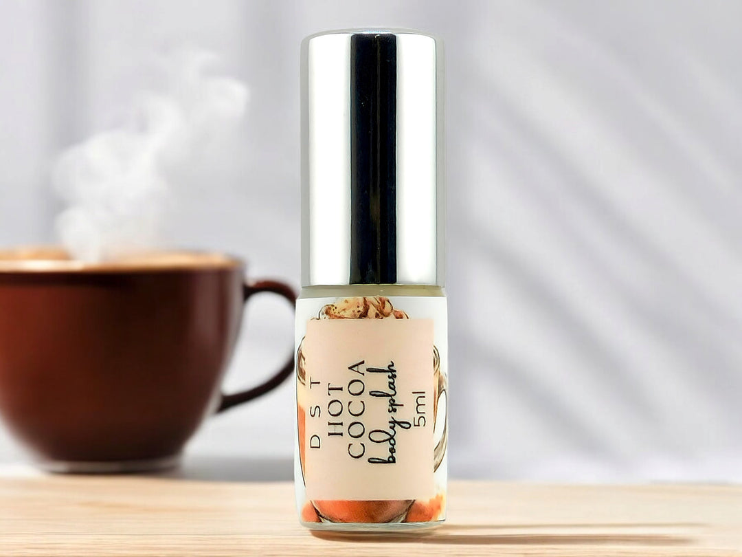 5ml bottle of Hot Cocoa body splash sitting on wood countertop with a steaming cup of coffee in the white shadowy background.