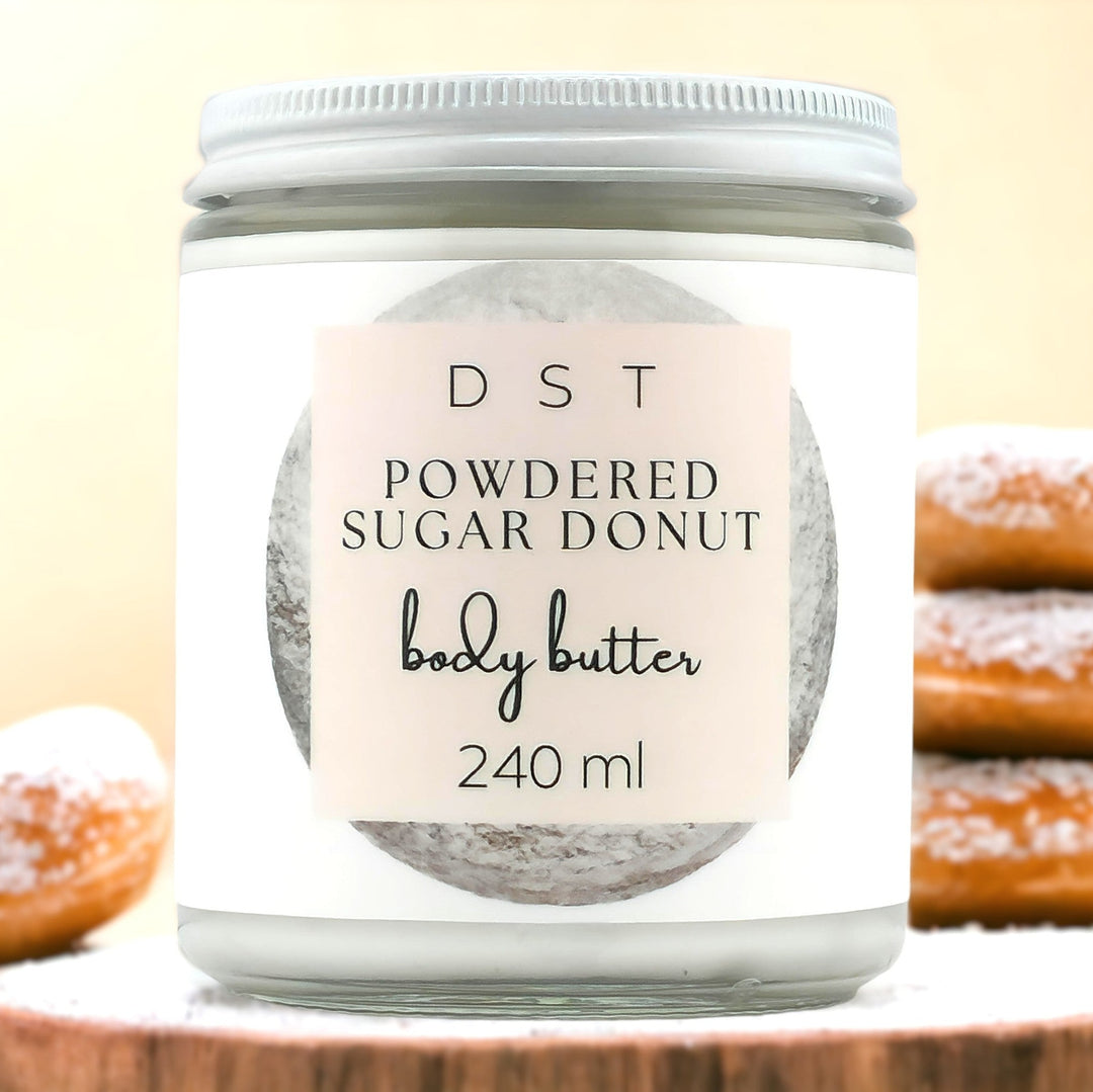 Eight ounce jar of powdered sugar donut scented body butter sitting on a wood countertop with powdered sugared doughnuts against a cream background.