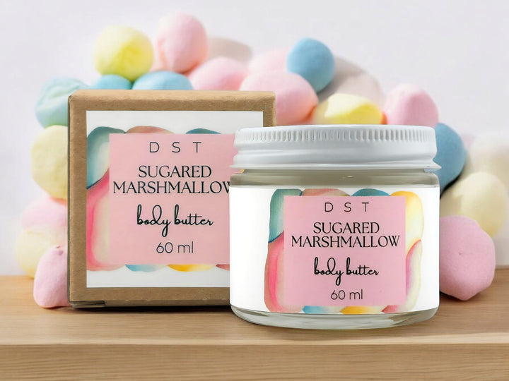 60ml jar of Sugared Marshmallow scented body butter and the box it is packaged in sitting on wood countertop with colorful pastel puffy marshmallows in the white background