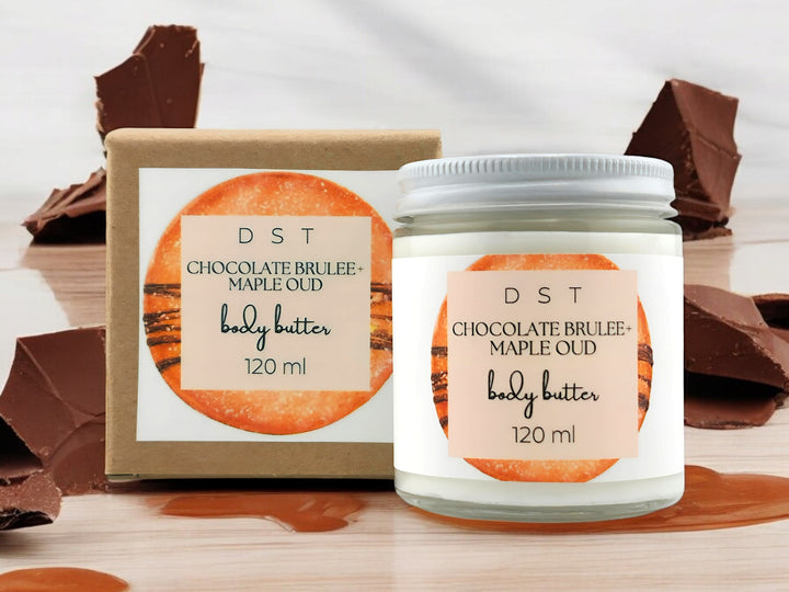 120ml jar of Chocolate Brulee and Maple Oud scented body butter and the box it is packaged in sitting on wood countertop with chocolate chunks and maple syrup drizzled in the white shadowy background.