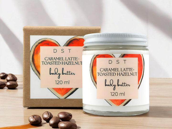 120ml jar of Caramel latte and toasted hazelnut scented body butter and the box it is packaged in sitting on wood countertop with coffee beans and caramel drizzled in the white shadowy background.