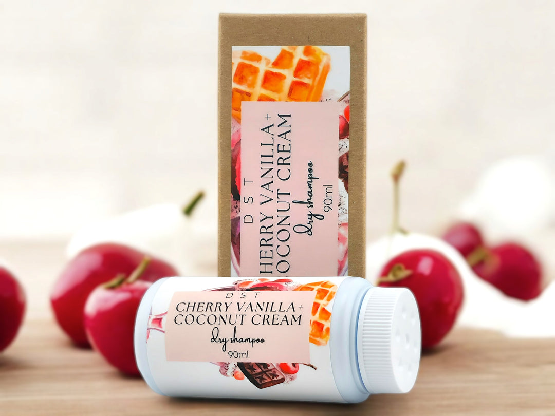 90ml bottle of Cherry Vanilla and Coconut Cream scented body powder and the box it is packaged in sitting on wood countertop with bright red cherries and whipped crem in the white background.